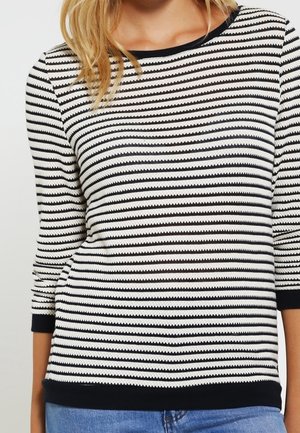 Mujer vistiendo un top de manga larga con rayas horizontales en blanco y negro, con puños y dobladillo negros, combinado con vaqueros azules.