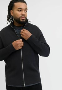 Czarna, żebrowana kurtka zapinana na zamek z wysokim kołnierzem, z metalowym zamkiem, noszona na ciemnoszarym T-shircie. Zbliżenie na ręce, które poprawiają zamek.