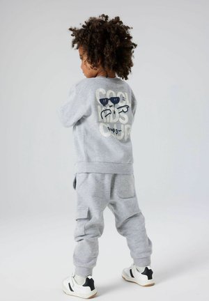 Ensemble de sweat-shirt et pantalon gris en coton. Le sweat-shirt présente un graphisme de lunettes de soleil et du texte dans le dos. Les chaussures sont blanches avec des accents noirs.