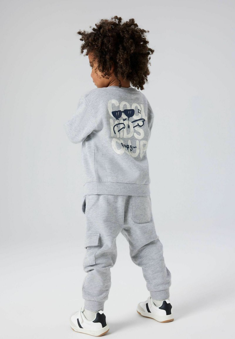 Conjunto de sudadera y pantalones grises de algodón. La sudadera presenta un gráfico de gafas de sol y texto en la parte posterior. Los zapatos son blancos con detalles en negro.
