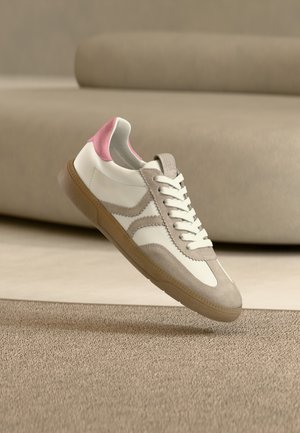 Baskets blanches avec des détails en suède beige, une touche rose au talon et une semelle en gomme, semblant flotter au-dessus d’un tapis brun clair dans une pièce minimaliste.