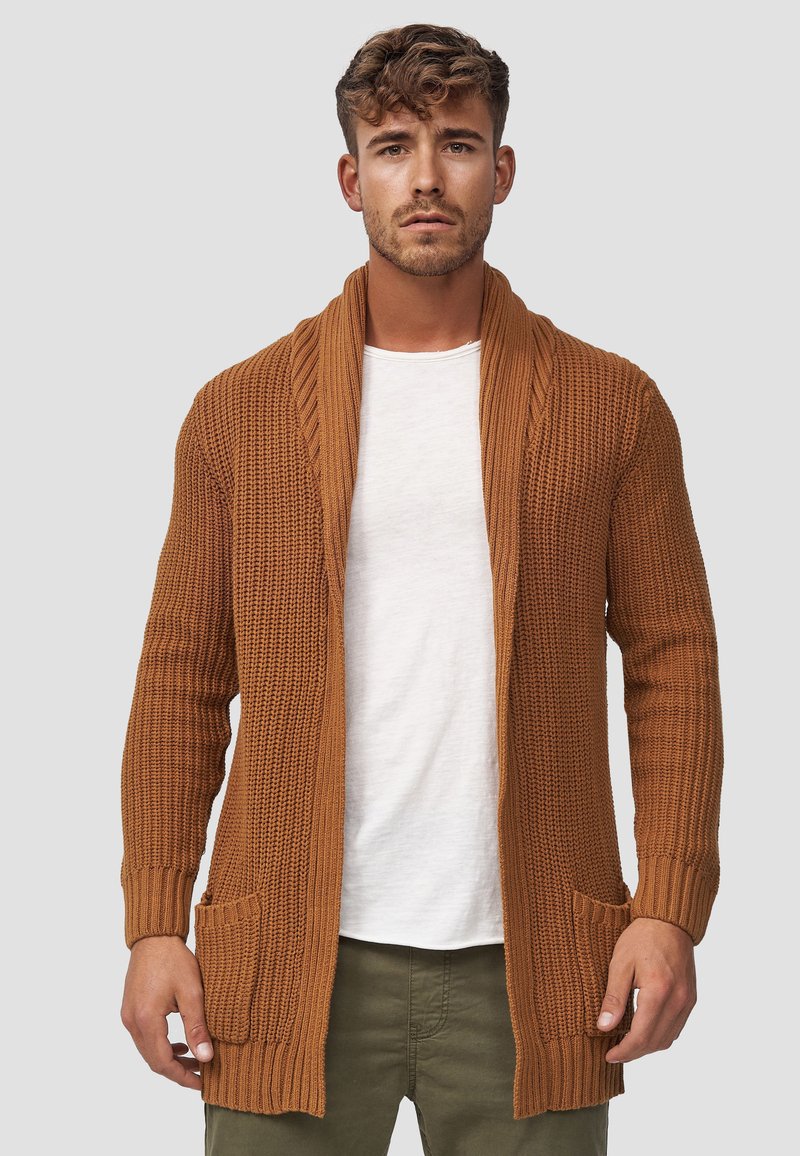 Brun strikket cardigan med sjalkrave, ribbet tekstur og to frontlommer, båret over en hvid skjorte. Casual, åben front design.
