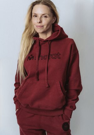 Vrouw met lang blond haar, gekleed in een bordeauxrode hoodie en bijpassende broek, staand met handen in de zakken tegen een lichte achtergrond.