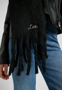 Écharpe en laine noire avec des extrémités frangées et un logo "Lee" brodé en rose. Portée avec une veste en cuir et un jean bleu.