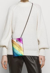 Barevná kovová taška přes rameno s diagonálními duhovými pruhy. Má řetízkový popruh a malý logový detail na vrchu.