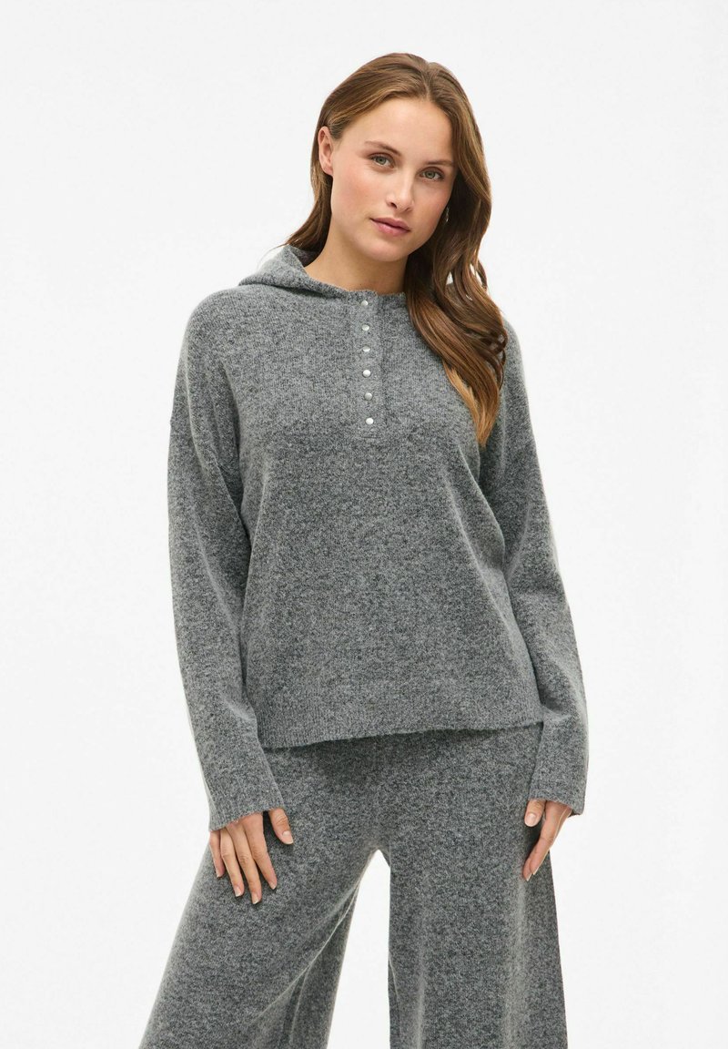 Pilkas gobtuvas megztinis su laisvu prigludimu, su mygtukais prie kaklo ir ilgomis rankovėmis, pagamintas iš minkštos tekstūruotos medžiagos.