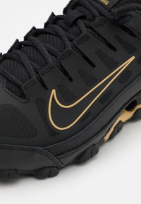 Sapatilha desportiva preta com uma parte superior em malha respirável, logótipo Swoosh dourado e sola de borracha texturizada com padrões de aderência visíveis.