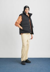 Μαύρο γιλέκο από πούπουλο με κλείσιμο κουμπιών και τσέπες, φορεμένο πάνω σε πορτοκαλί φούτερ. Μοντέλο σε καφέ παντελόνια και μαύρα παπούτσια σε μπλε χαλί.