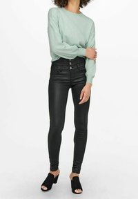 Persona con un top de manga larga verde claro, pantalones negros ajustados de cintura alta con botones y sandalias de tacón abierto negras, de pie.