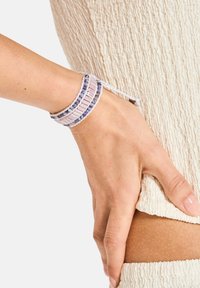 Monivärinen helmiranneketju, jossa on violetteja ja vaaleanpunaisia sävyjä, erityisesti punottu muotoilu ja liukuva suljin, kiedottuna ranteeseen tekstuurisen kankaan päällä.