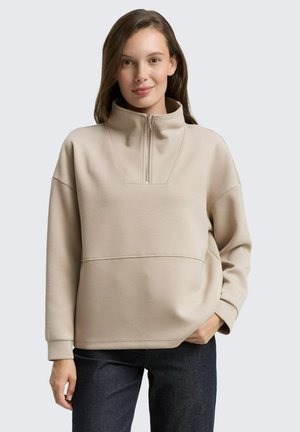 Pull beige avec col haut et demi-fermeture éclair. Fabriqué dans un matériau doux, avec des épaules tombantes et une couture sur la poitrine.