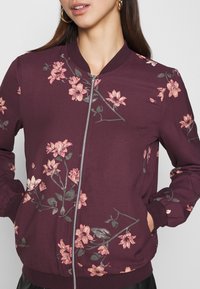 Burgundská bomber bunda s květinovým vzorem v růžové a zelené barvě. Má přední zip a žebrované manžety, vyrobena z hladkého materiálu.