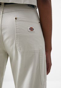 Bež pamučne hlače s stražnjim džepom, s ušivenom oznakom Dickies, metalnim zakovicama i opuštenim krojem.