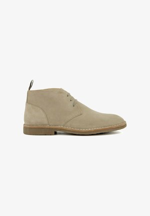 Bottine désert en suède beige avec trois œillets, couture de vissage, tirant au talon et semelle en caoutchouc marron clair sur fond blanc.