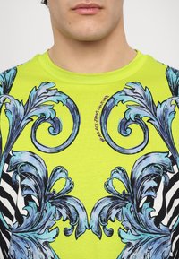 Limetkově zelené bavlněné tričko s modro-černým barokním vzorem se střídajícími pruhy zebry; obsahuje detail s textem "Versace Jeans Couture".