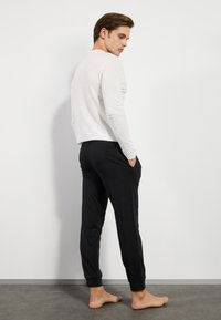 Langærmet hvid skjorte parret med sorte joggers med sidelommer og elastiske cuff; modellen står barfodet med en neutral baggrund.