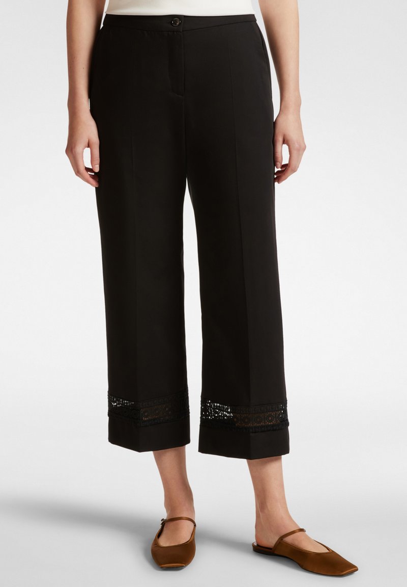 Pantaloni neri a gamba larga con una finitura liscia, caratterizzati da un bordo in pizzo all'orlo. Chiusura con bottone in vita. Abbinati a ballerine marroni.