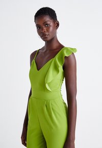 Žalias šokinėjimo kostiumas su V formos iškirpte, viena raukšlėta petneša, prigludusia liemene ir klostuotais akcentais. Audinys turi glotnų, matinį tekstūrą.