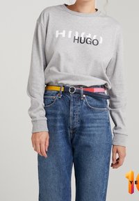 Grijze trui met de tekst "HUGO", gecombineerd met een blauwe spijkerbroek en een multicolor riem (geel, roze en blauw) met een zilveren gesp.