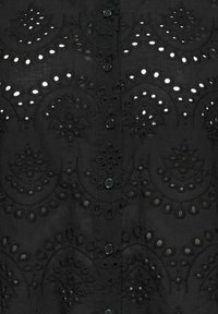 Zwart geborduurd shirt met ingewikkelde uitgesneden patronen, knoopsluitingen en textuurstof die een gelaagd ontwerp creëert.