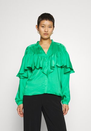 Femme portant un chemisier en satin vert vif avec des détails volants sur la poitrine et un pantalon noir, se tenant devant un fond uni.