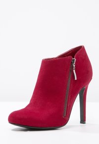 Bottine en daim rouge avec un bout effilé, un détail de fermeture éclair latérale et un talon aiguille haut. Texture lisse avec une finition mate.