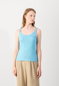 Light blue sleeveless top with textured fabric, featuring decorative loops on the straps. Paired with beige, pleated trousers.