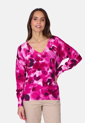 Femme portant un pull à col en V à fleurs rose et un pantalon beige, souriant avec une main sur la hanche sur un fond blanc uni.