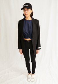 Blazer negro, camisa anudada azul marino, jeans skinny negros, zapatillas blancas de slip-on con textura perforada y una gorra negra con letras blancas.