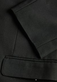 Black tailored blazer with a smooth texture, featuring a structured collar and neat stitching along the edges of the lapel and pocket.