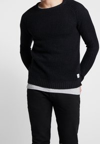 Czarny sweter robiony na drutach o prążkowanej fakturze, okrągłym dekolcie i długich rękawach, zestawiony z szarym podkoszulkiem i czarnymi spodniami.