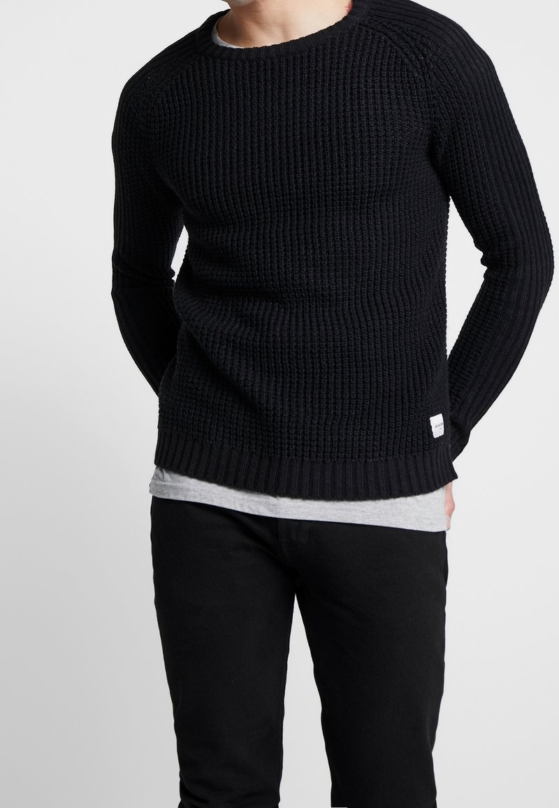 Czarny sweter robiony na drutach o prążkowanej fakturze, okrągłym dekolcie i długich rękawach, zestawiony z szarym podkoszulkiem i czarnymi spodniami.