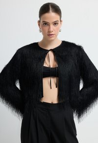 Kobieta w czarnej, fakturowej krótkiej kurtce noszonej na czarnym topie bandeau oraz czarnych cygaretkach, stojąca na tle białym.