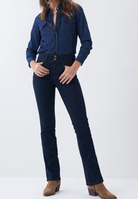 Chemise en denim bleu profond avec deux poches poitrine, des manches longues et une fermeture à boutons sur le devant. Associée à un jean évasé bleu foncé et des bottines brunes.