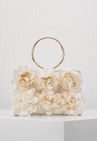 Sac à main blanc avec une anse circulaire dorée, orné de fleurs en tissu couleur crème, présentant des pétales superposés et un design structuré.
