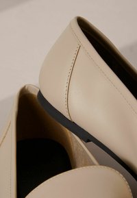 Mocassini in pelle beige con finitura liscia, suole in gomma nere basse e dettagli di cucitura in contrasto lungo le cuciture.