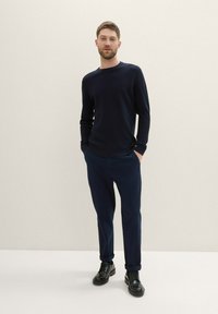 Navy gebreide trui met textuur, gecombineerd met een navy-tailored broek en zwarte veterschoenen. Model staat met de handen in de zakken tegen een lichte achtergrond.