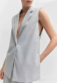 Bezrukávový svetle šedý blejzr s jedním knoflíkem, hlubokým V výstřihem a uvolněným střihem, nošený osobou, která má jednu ruku v kapse.