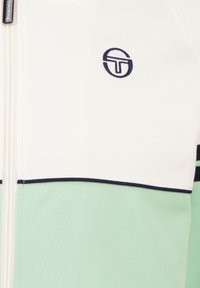 Glattjakke i hvit og mintgrønn. Har marineblå kant og logo. Glatt tekstur, atletisk design og mykt stoff.