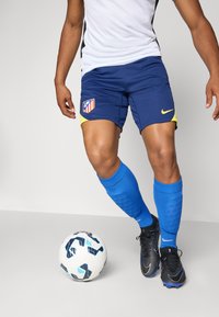 Nike fodboldshorts i marineblå med gule accenter og Atlético Madrid-logo; modellen har blå sokker og sorte fodboldstøvler på, stående ved en mønstret bold.
