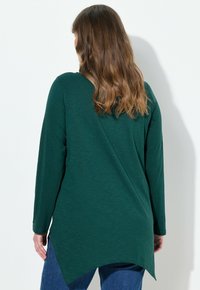 Grünes langärmeliges Oberteil mit einem gebogenen Hi-Low-Saum. Weicher, strukturierter Stoff und lässige Passform. Getragen mit blauen Jeans.