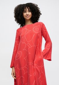 Frau mit lockigem Haar, die ein langärmeliges rotes Kleid mit großen hellrosa Blumenmustern trägt, vor einem schlichten weißen Hintergrund stehend.