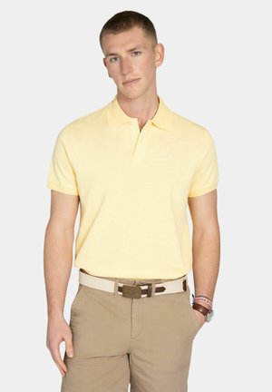 Joven con una camisa tipo polo amarilla clara, pantalones beige con un cinturón blanco, reloj de pulsera y una pulsera rosa sobre un fondo liso.