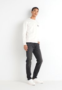Pullover bianco a collo rotondo con un piccolo logo, abbinato a jeans scuri e sneakers bianche. Il modello si trova su uno sfondo neutro, con le mani rilassate.