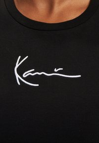 Black ribbed t-shirt featuring white embroidered signature across the chest. Crew neck design, close-fitting with a smooth texture.