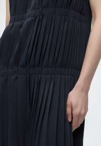 Gros plan d'une personne portant une robe noire plissée, montrant la texture détaillée du tissu et la main droite reposant à côté du corps.
