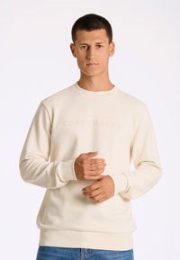 Ljust beige sweatshirt med rund hals, texturerat tyg och upphöjd "COPENHAGEN"-logotyp på framsidan. Har ribbade muddar och nederkant.