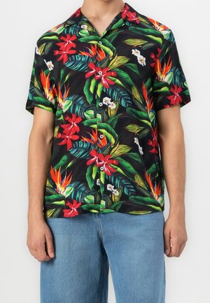 Mand iført sort kortærmet skjorte med farverigt tropisk blomsterprint og blå jeans, stående mod en ensfarvet baggrund.