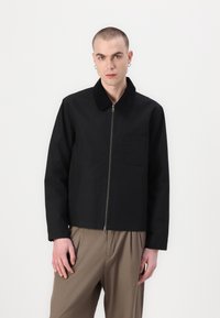 Chaqueta de algodón negra con cierre de cremallera, cuello en contraste y un bolsillo en el pecho; el modelo lleva puestos pantalones holgados beige.