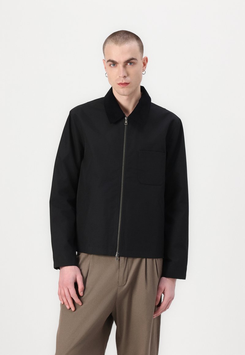 Chaqueta de algodón negra con cierre de cremallera, cuello en contraste y un bolsillo en el pecho; el modelo lleva puestos pantalones holgados beige.